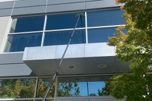 Commercial Window Cleaning Bay Area - Transparent Window Washing A long window-cleaning pole sprays water on a large glass office window of a gray building, with water streaks on the pane and trees reflected nearby.