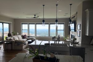 Open-concept living room and kitchen with a large window view of hills; white sofa, island, and pendant lights in a sunlit space.