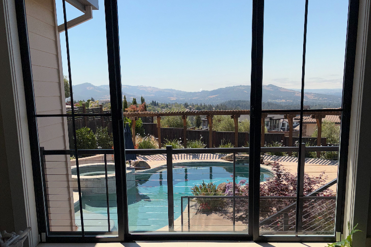 View from a glass-door interior looking out to a backyard pool, wooden deck, and distant mountains.