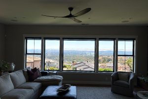 Bright living room with a panoramic wall of windows showing a hillside landscape and distant suburbs; beige sectional sofa and gray chair nearby.