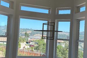 Bright room with tall rectangular windows and a geometric pendant light, overlooking a sunny hillside neighborhood.