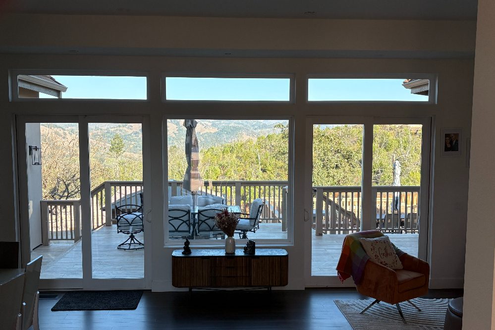 Interior living room with a glass door wall opening to a sunny wooden deck and scenic hillside view beyond the trees.