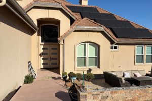 Beige stucco house with solar panels on a sloped roof, a arched window and beige front door, and a patio with outdoor seating.