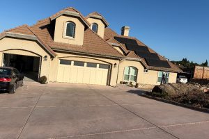Beige stucco house with brown tile roof and solar panels on the right wing, large concrete driveway and closed garage doors.
