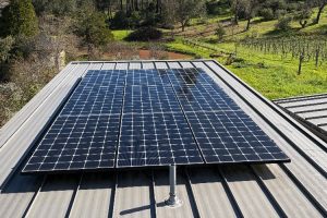 Solar panels installed on a metal rooftop with a rural field and trees in the background.