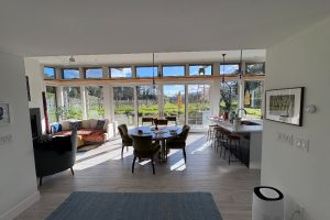 Open-plan living/dining/kitchen area with large glass walls looking out onto a vineyard; round dining table with chairs and a modern island with bar stools nearby.