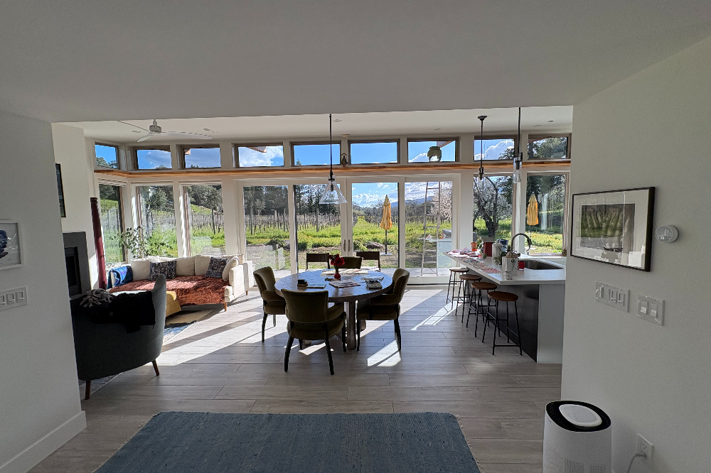 Open-plan living/dining/kitchen area with large glass walls looking out onto a vineyard; round dining table with chairs and a modern island with bar stools nearby.
