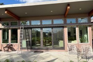 Patio of a modern home with large glass doors, a ladder leaning against the door, and white wicker seating under a blue sky.