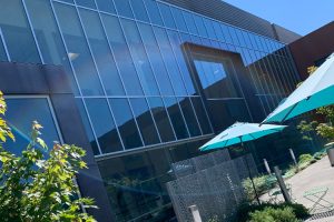 Modern glass-front building with blue-tinted windows, an outdoor patio area, and teal umbrellas casting shade on metal tables and chairs.