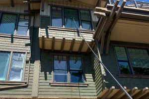 Person uses a long cleaning pole to wash exterior windows on a multi-story green building, reaching the upper balcony area.