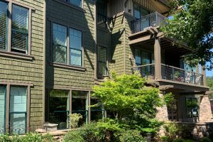 Green-siding apartment building with large windows, a wooden balcony, and a landscaped front yard.