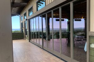Covered wooden terrace with floor-to-ceiling glass doors; patio seating visible inside overlooking a distant landscape on a sunny day.