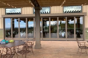 Outdoor patio with round metal tables and chairs in front of a glass-walled restaurant entrance; interior chandeliers visible through the doors.