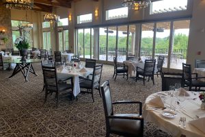Restaurant dining room with white-tablecloth tables set for service and large windows opening to an outdoor patio outside.