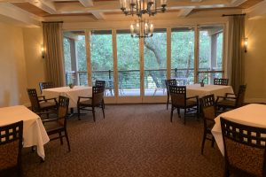 Cozy restaurant dining room with white-tablecloth tables, brown chairs, and a glass-door balcony overlooking trees outside.