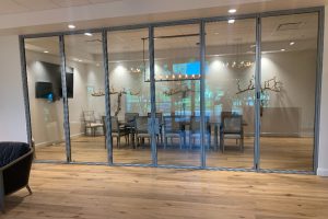 Glass-walled conference room with a long table and chairs, seen through sliding doors, wood floor and bare-tree decor under a chandelier.