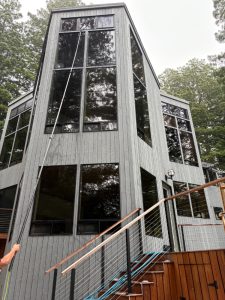 Tall gray wooden house with large windows in a wooded setting; a window-cleaning pole reaches the upper pane.
