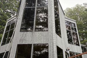 Modern gray, timber-clad house with large floor-to-ceiling windows reflecting surrounding trees in a woodland setting, viewed from the corner.