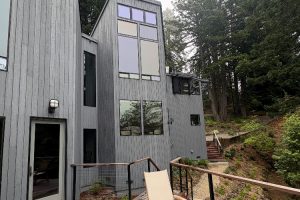 Modern gray wooden house with large multi-pane windows on a hillside, a deck with a chair, and surrounding trees.