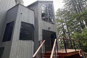 Modern gray wooden house with large angular windows and a wooden deck, set among trees outside. A metal railing runs along the deck stairs to the entrance.