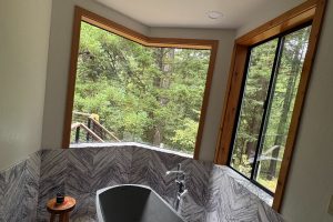 Freestanding oval bathtub in a marble-tiled bathroom with large angled windows overlooking a forested yard and wooden trim