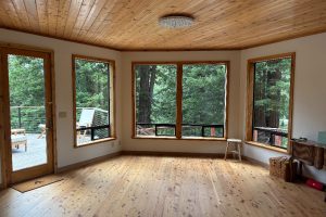 A sunlit, empty wooden room with large corner windows showing a forest; deck with chairs outside.