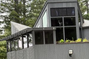 Modern gray wooden house with a wraparound sunroom and tall, dark-framed windows set among trees on a cloudy day.