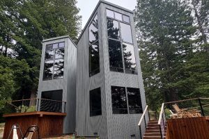 Modern gray wooden house with tall, angular windows set among trees, a wooden deck on the left and stairs on the right leading to outdoor seating.