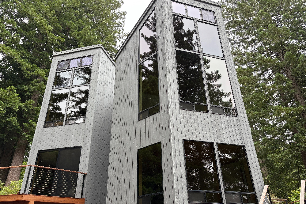 Tall, modern gray metal house with large floor-to-ceiling windows surrounded by trees in a wooded setting.