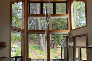 Tall wooden-framed window filling the interior, opening to a forested yard with tall trees; a glass wind-chime hangs in the center.