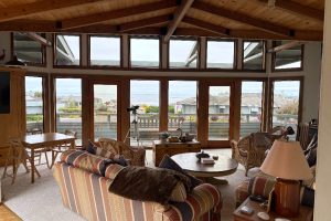 Cozy living room with striped sofa, wicker chairs, and a view of the ocean through tall glass doors and windows.