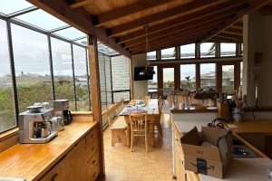 Bright open-plan kitchen and dining area with exposed wooden beams, a coffee station on a wooden counter, and a long dining table beneath skylights in daylight.
