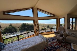 Panoramic bedroom with large angled windows showing a grassy hillside and blue sky; a bed, recliner, and sofa with an 'Oyster Catcher' pillow nearby.