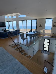 IMG_0466 - Transparent Window Washing Open-concept living and dining area with glass railing overlooking a beach and ocean through large floor-to-ceiling windows.