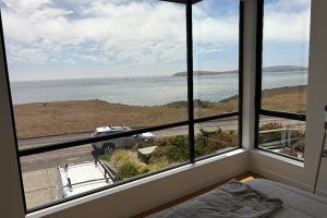 View from a bedroom window overlooking the ocean and rocky coastline, with a car parked outside and a bed in the foreground comforter visible.