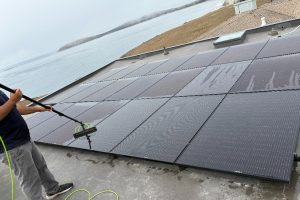 Person on a flat roof cleaning rows of solar panels with a long-handled roller attachment on a hose nearby.