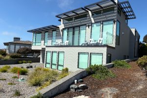 Modern two-story house with large glass windows and a glass-railed balcony, set on a desert landscape under a bright blue sky