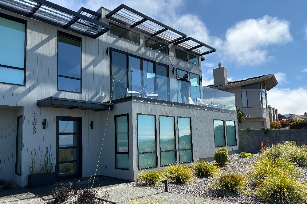 Modern two-story house with glass balcony, large blue-tinted windows, and a desert-inspired front yard.