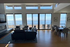 Bright, modern living room with floor-to-ceiling windows overlooking the ocean; sofa, chairs, and a dining table nearby on a wooden floor.