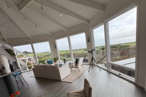 Bright living room with floor-to-ceiling windows showing a coastal landscape, white sofa and armchairs, and a tripod-mounted telescope nearby.