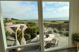 Indoor view through a large window to a coastal patio with a white dining table and chairs, manicured shrubs, and the sea on the horizon.