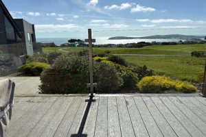 Deck with glass railing facing a coastal landscape; a wooden cross stands at the center with a garden and the sea beyond.