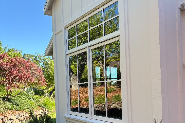 White-framed window with grid panes on a sunny house, reflecting a green garden and blue sky outside.