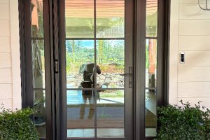 IMG_1987 - Transparent Window Washing Double glass doors with dark frames, reflecting a person taking a photo, opening to a sunny patio beyond with a stone wall and greenery in view.