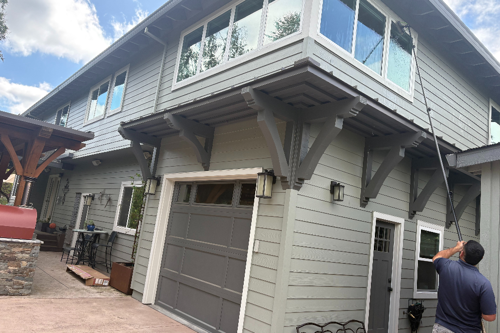 Man on the right uses a long pole to reach high windows on a gray two-story house with a large garage door, outdoors on a sunny day.] ,