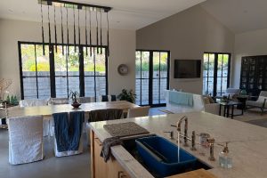 Open-concept kitchen and living area with a marble island sink and a blue dish tub, large windows, and a dining table with white covers.