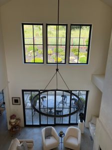 Overhead view of a bright living room: two white armchairs around a small table, large circular chandelier, and floor-to-ceiling windows opening to a patio and garden outside.