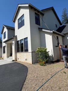 Two-story modern house with light siding; a worker uses an extendable pole to clean upper-story windows on the right.