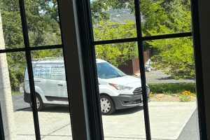 View through window panes of a white van labeled 'Transparent Window Washing' parked in a driveway with trees and a garden in the background.