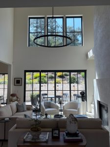 Two-story living room with a large circular pendant chandelier, black-framed windows, and a glass door patio beyond. Neutral seating and plants fill the space.